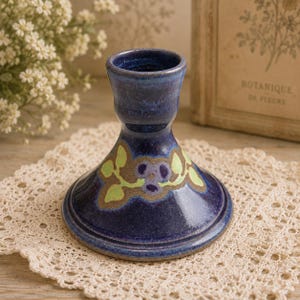 May include: A dark blue ceramic candle holder with a wide, flared base and a cylindrical top. The surface features a hand-painted design of light green leaves and a central purple flower motif set against a textured, earthy brown background.