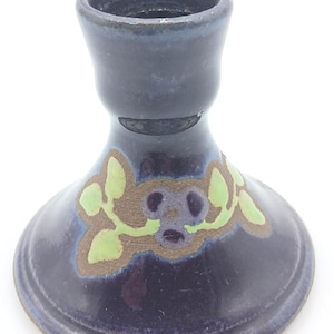 May include: A dark blue ceramic candle holder with a glossy finish and a wide, flared base. The exterior features a hand-painted decorative motif consisting of light green leaves and a central dark blue flower shape outlined in a muted brown tone.