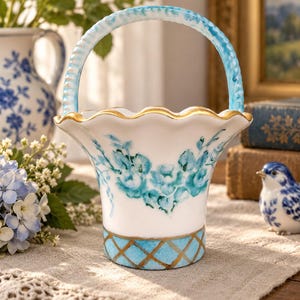 May include: A white ceramic basket with a scalloped gold rim and a curved handle. The exterior features hand-painted blue floral motifs and a blue and gold lattice pattern at the base. The basket has a glossy finish and a wide, flared opening.