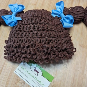 May include: A brown crocheted baby hat with attached yarn hair and two blue satin bows. The hat is handmade and has a textured appearance. The hat is designed to look like a child's hairstyle.