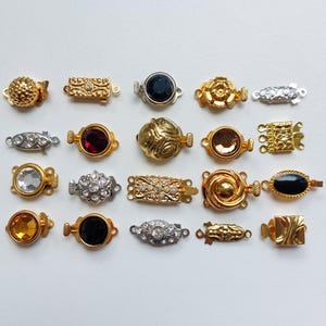 Puede incluir: Surtido de 18 cierres de metal dorado, plateado y negro para collares y pulseras. Los cierres presentan diversos diseños, incluyendo formas redondas, ovaladas y rectangulares, algunos adornados con pedrería o piedras de colores.