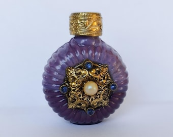 May include: A round, purple glass perfume bottle with a ribbed texture and a gold-toned metal cap. The center features an ornate gold-toned metal filigree emblem decorated with a central white pearl and four small blue stones at the points.