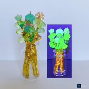 Può includere: Un piccolo vaso di vetro pieno di perline color oro e sormontato da un bouquet di ciondoli a forma di fiore in vetro verde e trasparente. I fiori hanno varie forme e dimensioni, con accenti dorati. Il vaso è trasparente e lo sfondo è chiaro.