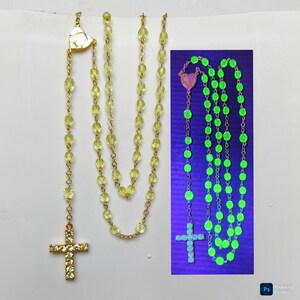 Puede incluir: Un rosario con cuentas amarillas y un colgante de cruz dorada. La cruz y el conector en forma de corazón están adornados con pequeñas piedras transparentes. Un segundo rosario brilla de color verde brillante bajo luz negra.