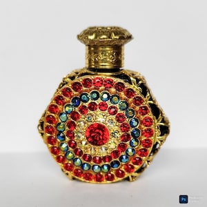 Può includere: Un flacone di profumo decorativo con un pannello frontale circolare ornato da anelli concentrici di strass rossi, blu e trasparenti. Il flacone presenta una cornice in metallo dorato con un tappo testurizzato e un design in filigrana.