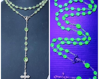 Pode incluir: Um colar de rosário de vidro verde com um pingente de cruz de prata. As contas brilham intensamente sob uma luz negra.