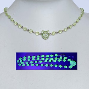 Peut inclure: Un collier avec un pendentif tête de chat en verre vert et une chaîne de petites perles en verre vert. Le collier brille en vert sous une lumière noire.