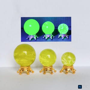 Op de afbeelding: Drie gele kristallen bollen van verschillende grootte, elk op een decoratieve goudkleurige standaard. Een kleinere inset-afbeelding toont dezelfde kristallen bollen groen oplichtend. De kristallen bollen hebben een troebel interieur.