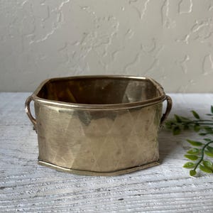 May include: A small, antique brass planter with a geometric pattern. The planter has a square shape with rounded corners and small handles on each side. The brass has a warm, golden hue and shows signs of age.
