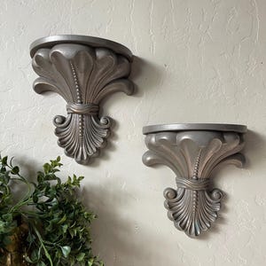 May include: Two decorative wall shelves with a silver finish. Each shelf has a rounded top and a detailed, stylized floral design. The shelves are mounted on a textured wall, offering a classic, elegant aesthetic for home decor.