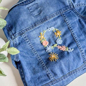 May include: A blue denim jacket with floral embroidery. The back of the jacket features a colorful embroidered floral design in yellow, pink, and blue, with the letter "R" at the center. The jacket is a light wash denim.