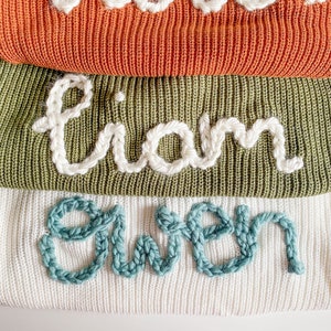 May include: Three knitted sweaters stacked on top of each other. The top sweater is orange, the middle sweater is green, and the bottom sweater is white. The sweaters have names embroidered on them: Liam, Gwen.