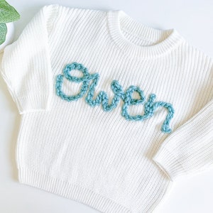 May include: White knitted sweater with the name "Owen" embroidered in teal blue yarn.