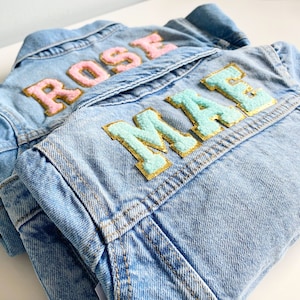 May include: Two denim jackets with personalized names, one with the name "ROSE" in pink letters and the other with the name "MAE" in light blue letters.