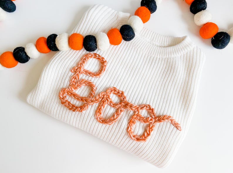 Op de afbeelding: Een witte trui met het woord "BOO" geborduurd in oranje garen. De trui is versierd met een guirlande van zwarte, oranje en witte pompoenen.