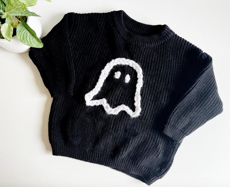 Puede incluir: Su&eacute;ter de punto negro con una aplicaci&oacute;n de fantasma blanco. El fantasma tiene dos ojos y una peque&ntilde;a sonrisa.