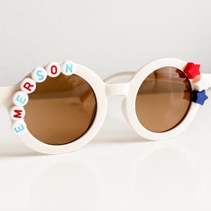 Puede incluir: Gafas de sol redondas blancas con lentes marrones. Las gafas tienen el nombre "EMERSON" escrito en letras de colores en el lado izquierdo y estrellas rojas, blancas y azules en el lado derecho.