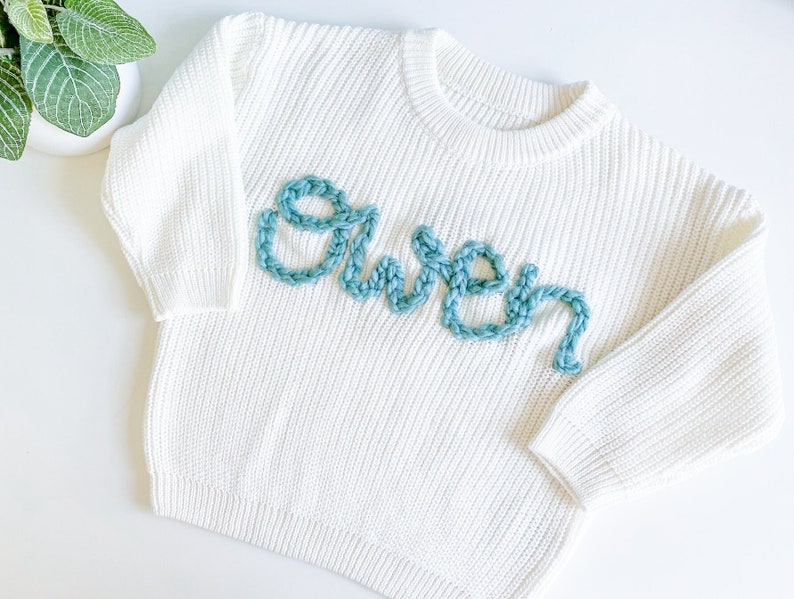 May include: White knitted sweater with the name "Owen" embroidered in teal blue yarn.