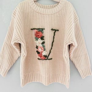 Puede incluir: Un suéter de punto color crema con una letra "V" bordada con flores en la parte delantera.