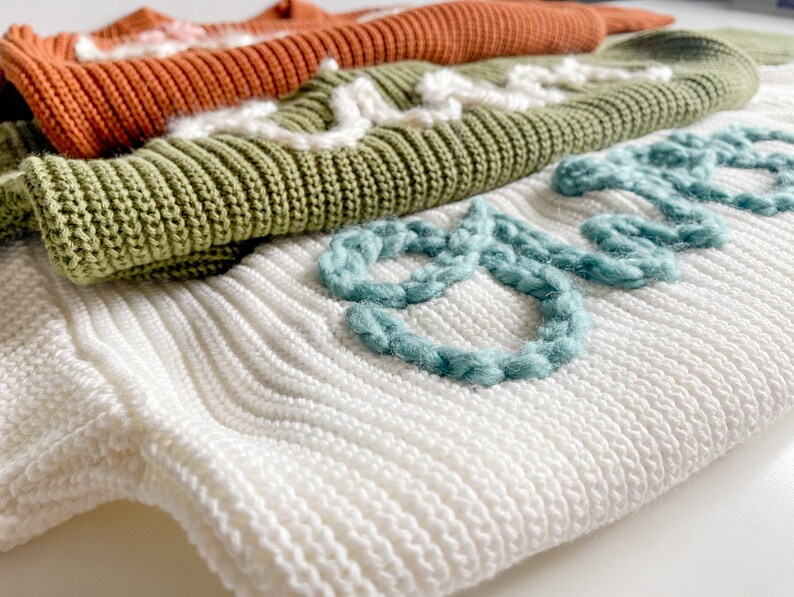 May include: A stack of knitted sweaters in white, green, and orange. The top sweater has the word "dream" embroidered in teal yarn.