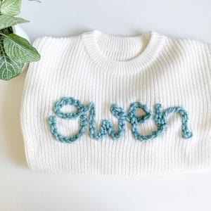 May include: White knitted sweater with the word "Gwen" embroidered in light blue yarn.