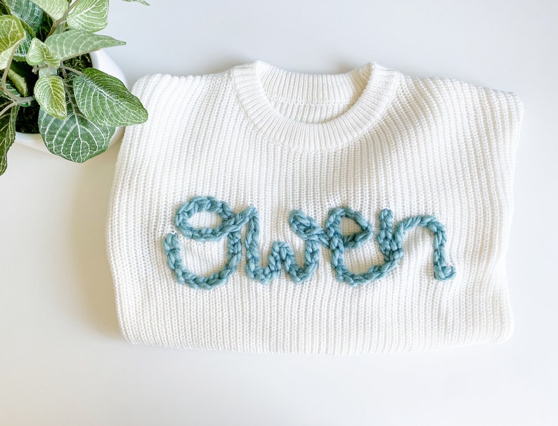 May include: White knitted sweater with the word "Gwen" embroidered in light blue yarn.