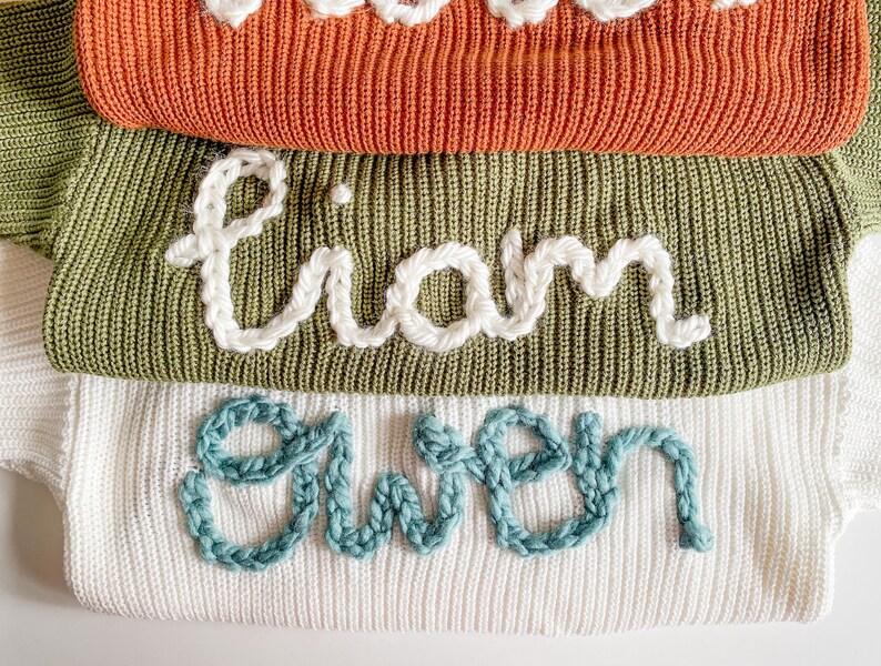 May include: Three knitted sweaters stacked on top of each other. The top sweater is orange, the middle sweater is green, and the bottom sweater is white. The sweaters have names embroidered on them: Liam, Gwen.