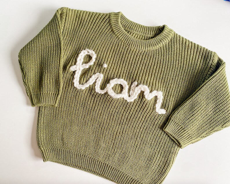 May include: A green knitted sweater with the name "Liam" embroidered in white yarn.