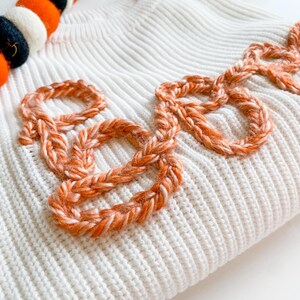 Op de afbeelding: Witte gebreide trui met het woord "Boo" geborduurd in oranje garen.