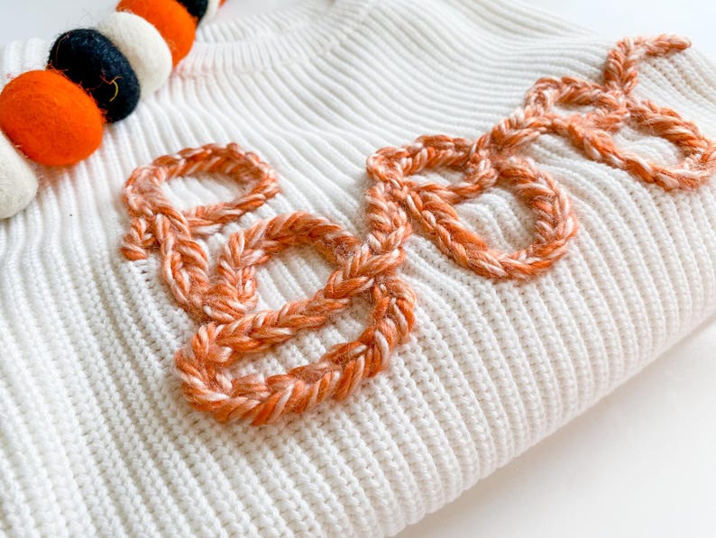 Op de afbeelding: Witte gebreide trui met het woord "Boo" geborduurd in oranje garen.