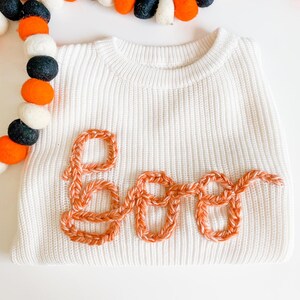 Op de afbeelding: Witte trui met een gehaakt oranje "BOO"-ontwerp.
