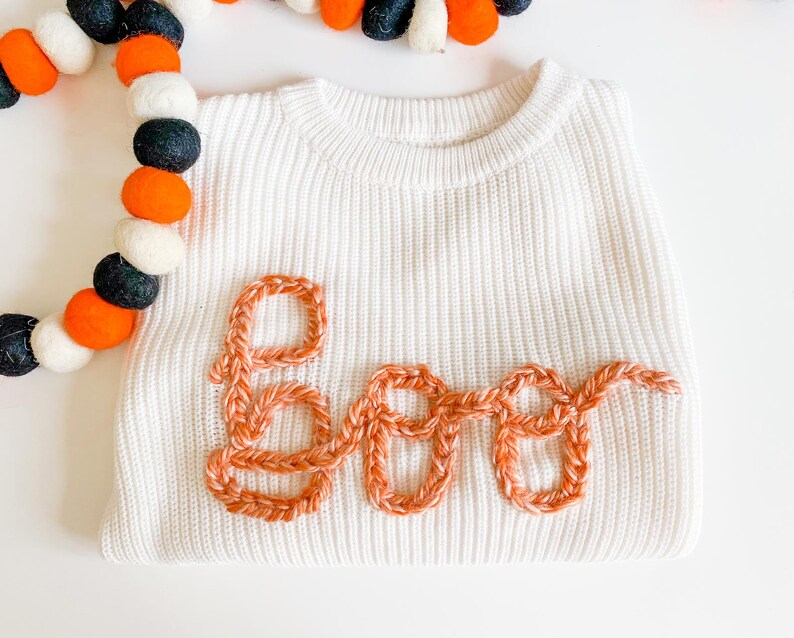 Op de afbeelding: Witte trui met een gehaakt oranje "BOO"-ontwerp.
