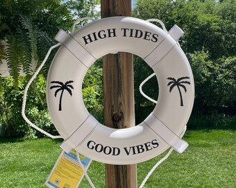 May include: White life preserver with black rope handles and black text that reads "HIGH TIDES GOOD VIBES". Two black palm trees are printed on the life preserver.
