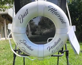 May include: A white life ring with black text that reads Just Married, Jordan & Shane, and Positano 10/12/22. The life ring features white fabric straps and a white rope threaded around the perimeter, providing a nautical appearance.