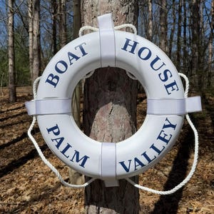 May include: White lifebuoy ring with the words "BOAT HOUSE PALM VALLEY" in navy blue lettering. The ring is mounted on a tree trunk with white straps and rope. The background shows a forest with brown leaves.