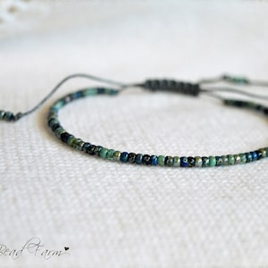 May include: A delicate bracelet made with tiny blue, green, and silver beads on a gray cord with a sliding knot closure.