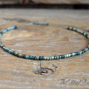 May include: A delicate beaded necklace with a sliding knot closure. The necklace features small, iridescent beads in shades of blue, green, and silver. The text "The Bead Farm" is visible in the lower right corner.