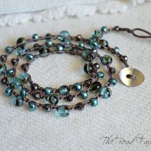 May include: A multi-strand necklace featuring teal and dark brown beads, strung on a brown cord. The necklace has a button closure and is displayed on a white, textured surface. The Bead Farm logo is visible in the bottom right corner.