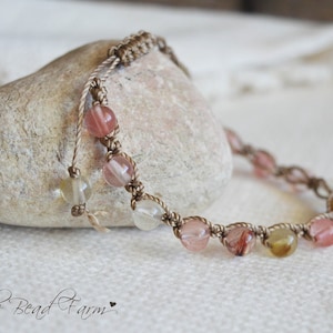 May include: A handmade macrame bracelet with clear and pink gemstone beads. The beads are strung on a beige cord, and the bracelet is displayed on a light-colored stone. The Bead Farm logo is visible in the bottom left corner.