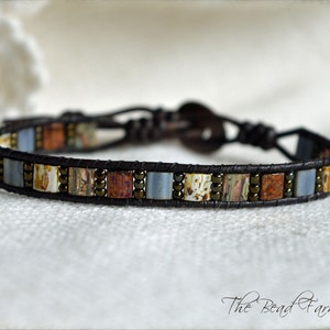 May include: A brown leather bracelet with a single row of colorful square beads. The beads are a mix of blue, white, brown, and orange.