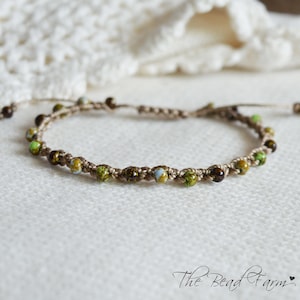 May include: A macrame bracelet crafted from tan cord featuring small, round, multi-colored beads in shades of green, brown, and light blue. The bracelet includes an adjustable sliding knot closure and the text The Bead Farm with a heart symbol.