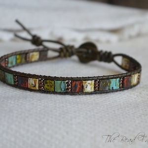 May include: A brown leather bracelet with a colorful square tile pattern. The bracelet has a brown leather cord with a knot closure.