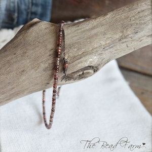 May include: A delicate bracelet made with small, iridescent brown and copper beads. The bracelet is strung on a thin, dark cord and is displayed on a piece of driftwood.