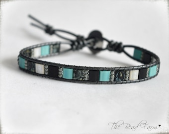May include: A wrap bracelet featuring a black leather band adorned with square beads in turquoise, black, white, and patterned gray. The bracelet has a button closure and adjustable leather ties. The Bead Farm logo is visible in the bottom right corner.