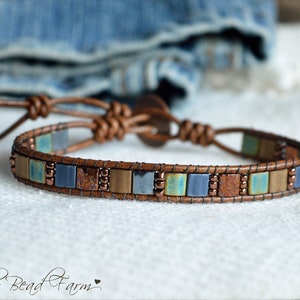May include: A brown leather bracelet with a row of square-shaped beads in shades of blue, green, and brown. The bracelet has a knotted closure.