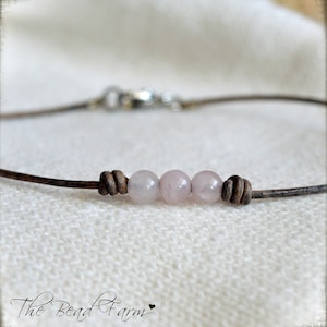 May include: A delicate bracelet featuring three pale pink rose quartz beads strung on a brown leather cord. The cord is knotted on each side of the beads and secured with a silver clasp. The Bead Farm logo is in the bottom left corner.