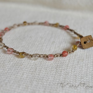May include: A delicate bracelet featuring a series of small, translucent pink, yellow, and clear beads knotted onto a thin, light brown cord. The bracelet is secured with a square, light-colored wooden button closure. The text The Bead Farm is visible.
