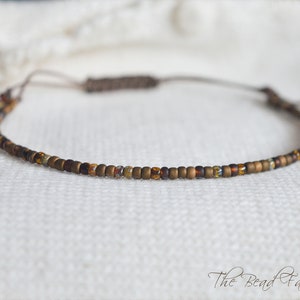 May include: A delicate brown and gold beaded bracelet with a simple adjustable cord closure. The bracelet is made with small, round beads and has a subtle shimmer.