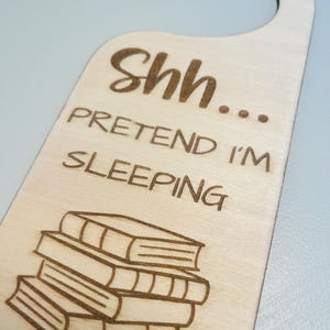 May include: Wooden door hanger with the text "Shh... Pretend I'm Sleeping" and a stack of books etched into the wood. The hanger has a curved top for easy hanging and a light wood grain finish.