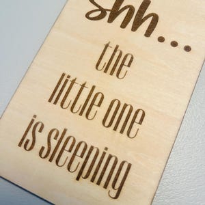 May include: A light-colored wooden sign with rounded edges. The sign has the words "Shh... the little one is sleeping" etched into the wood in a dark brown color. The sign is designed to be hung on a door.